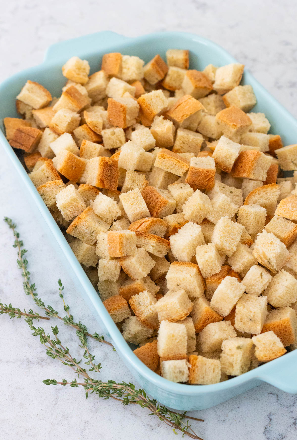 The cubed bread is in a blue baking dish next to a sprig of fresh thyme.