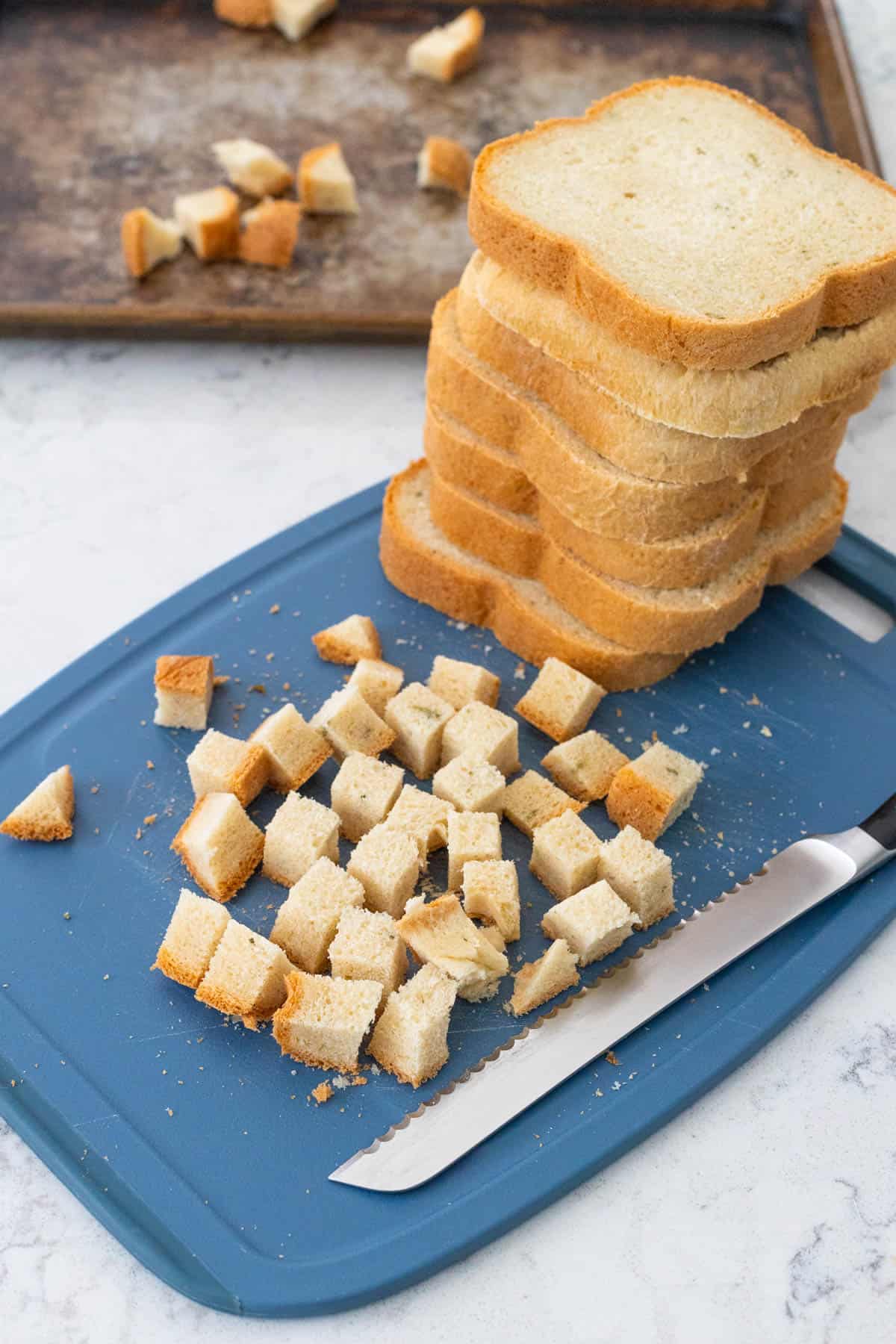The loaf of bread has been sliced and stacked, the individual slices are cubed on the cutting board.
