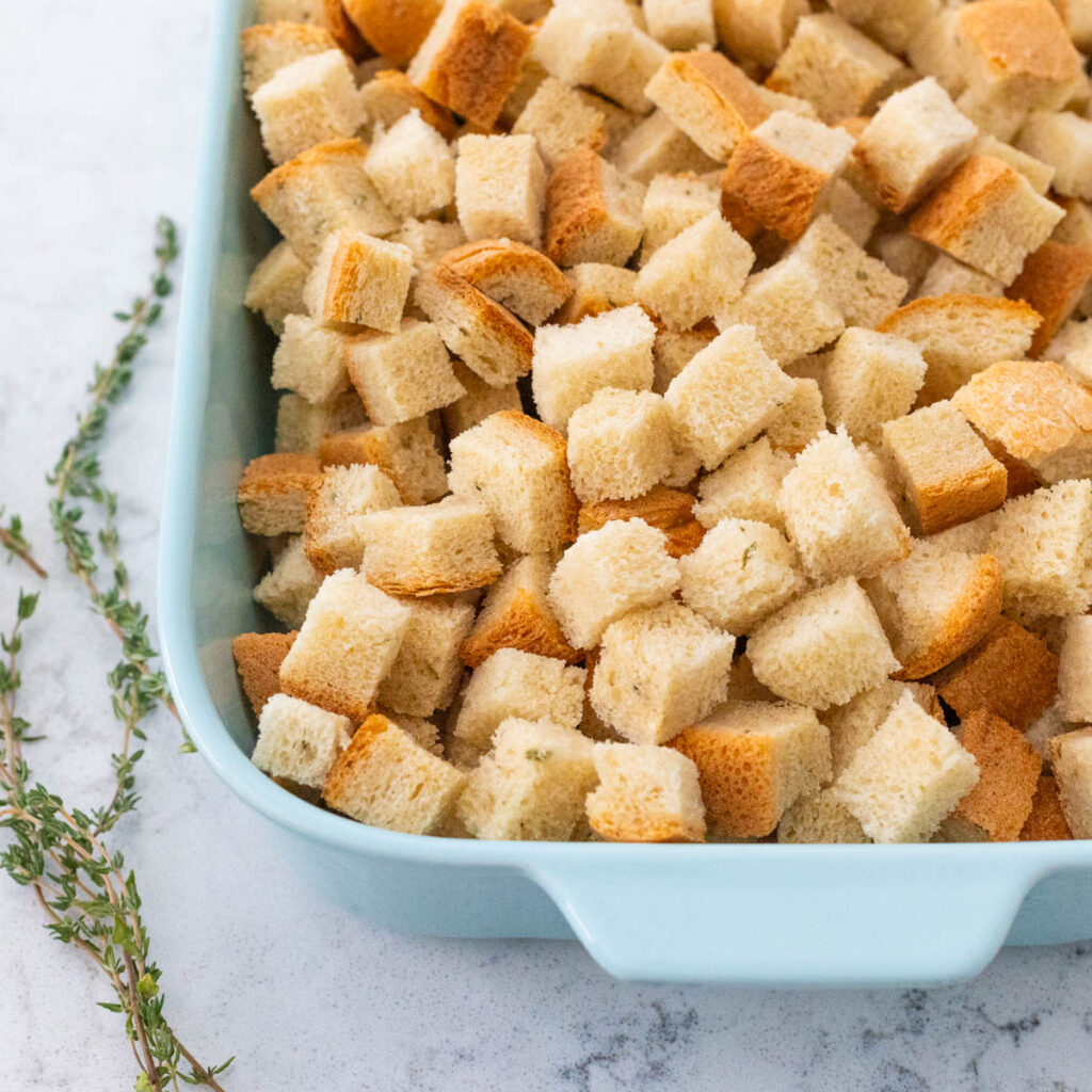 The cubed bread is in a baking dish next to a sprig of fresh thyme.