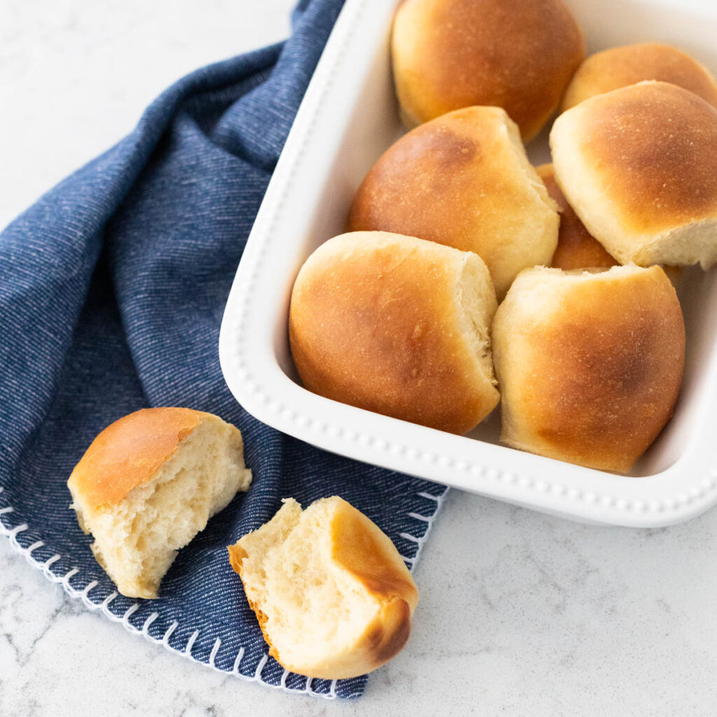 A white dish is filled with golden brown buttery dinner rolls. One roll has been broken open and rests on a blue napkin.