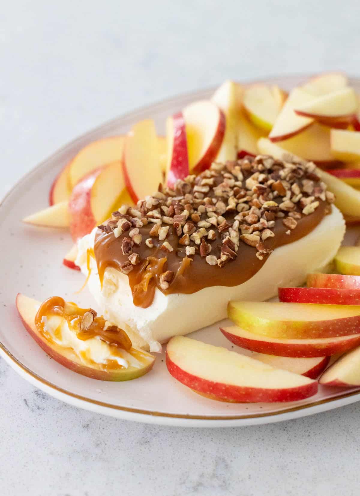 A brick of cream cheese has caramel and chopped pecans on top. Sliced apples are all around it on the platter.