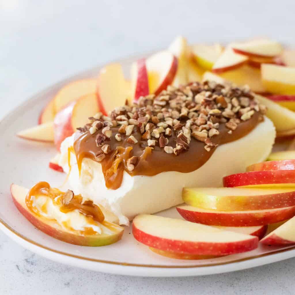 A brick of cream cheese has caramel and chopped pecans on top. Sliced apples are all around it on the platter.