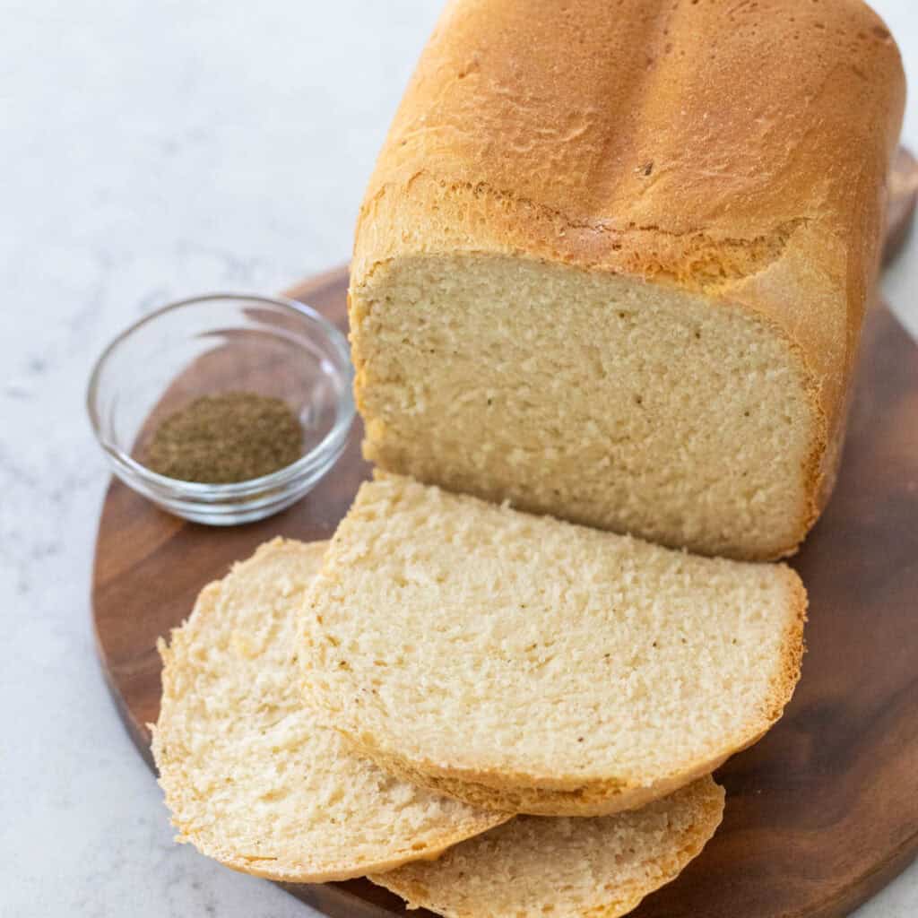 The loaf of celery seed bread has been sliced and sits on a board next to a cup of celery seeds.