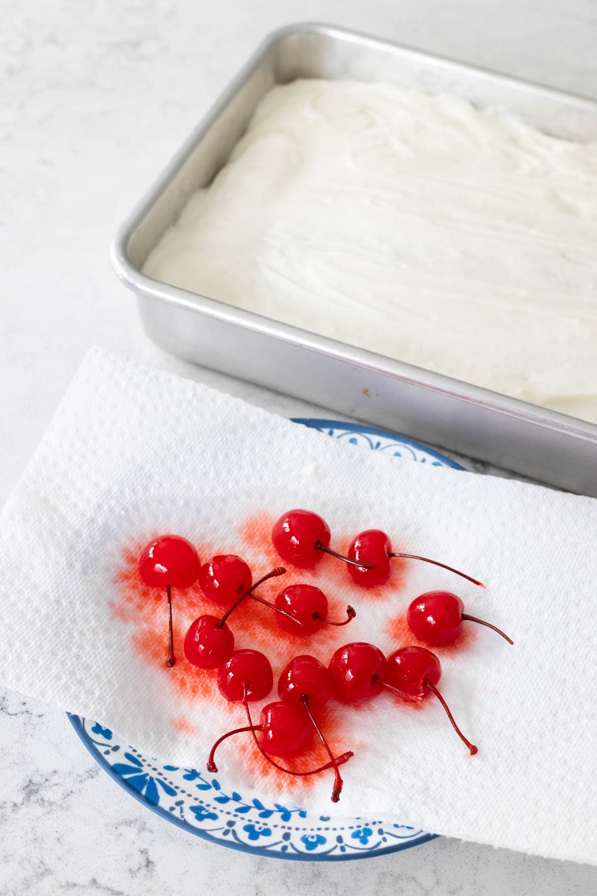 Some paper towel lines a small plate, the maraschino cherries are draining their juices on top. The iced cake waits in the back.