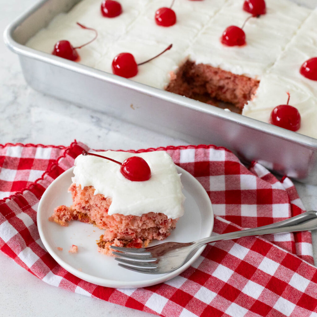 A slice of cherry cake is on a plate with a fork beside it. In the back is the metal cake pan the 9x13-inch cake was baked in. You can see the slice has been removed and there are maraschino cherries on top of the cake.