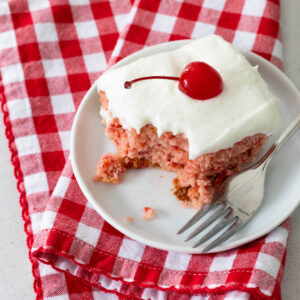 A square of pink cherry cake has white almond icing on the top and a maraschino cherry with a curved stem as garnish on top. The plate rests on a red and white checked napkin, a bite has been taken out of the cake.