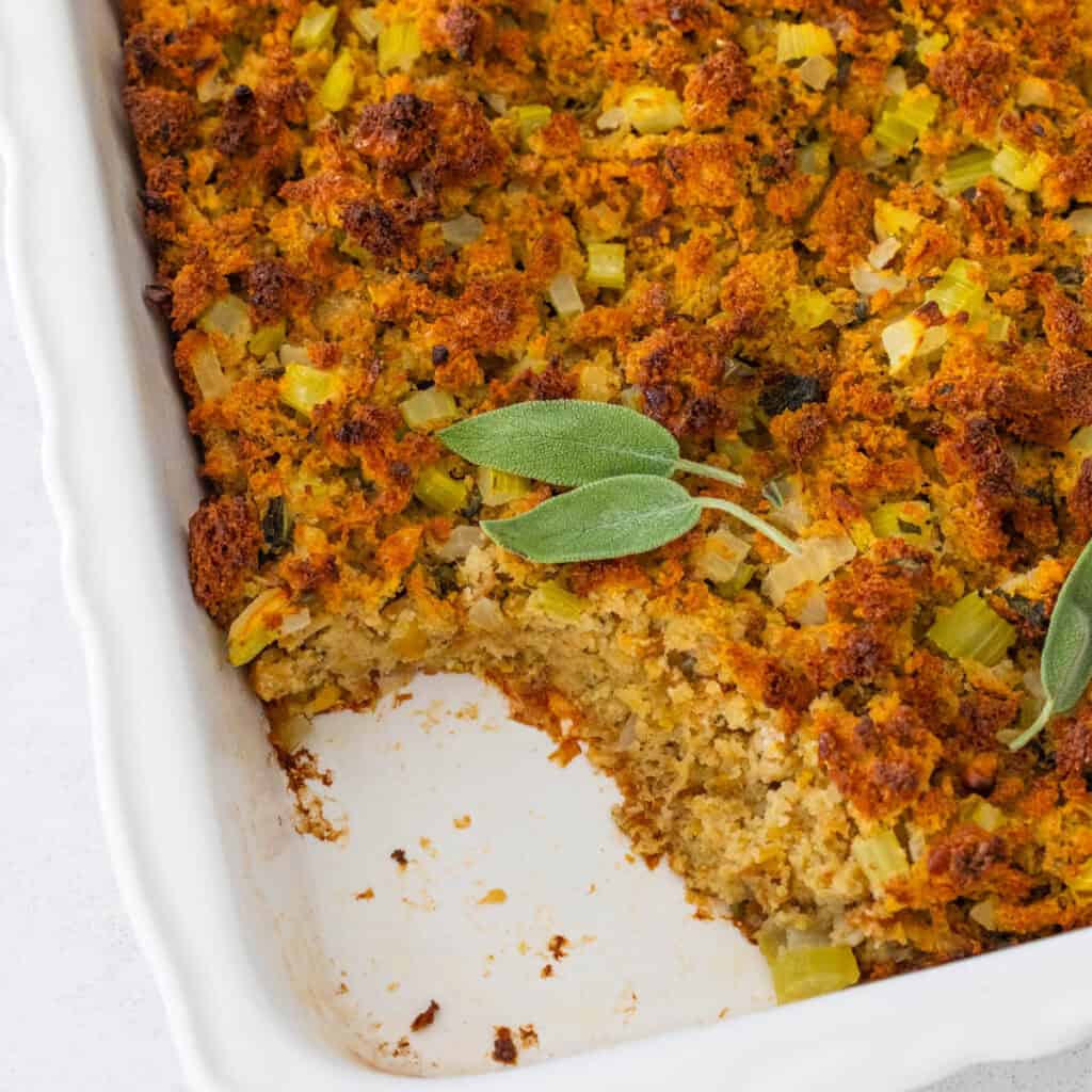 A baking dish with cornbread dressing in it has a serving scooped out. Fresh sage leaves are on top.