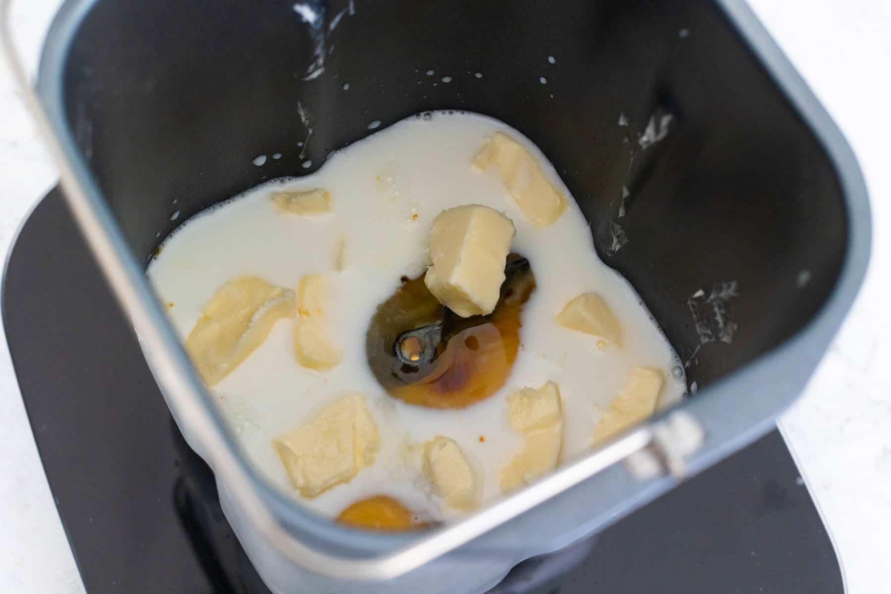 A bread maker pan filled with milk, butter pieces, an egg, and molasses ready to be mixed for gingerbread bread.
