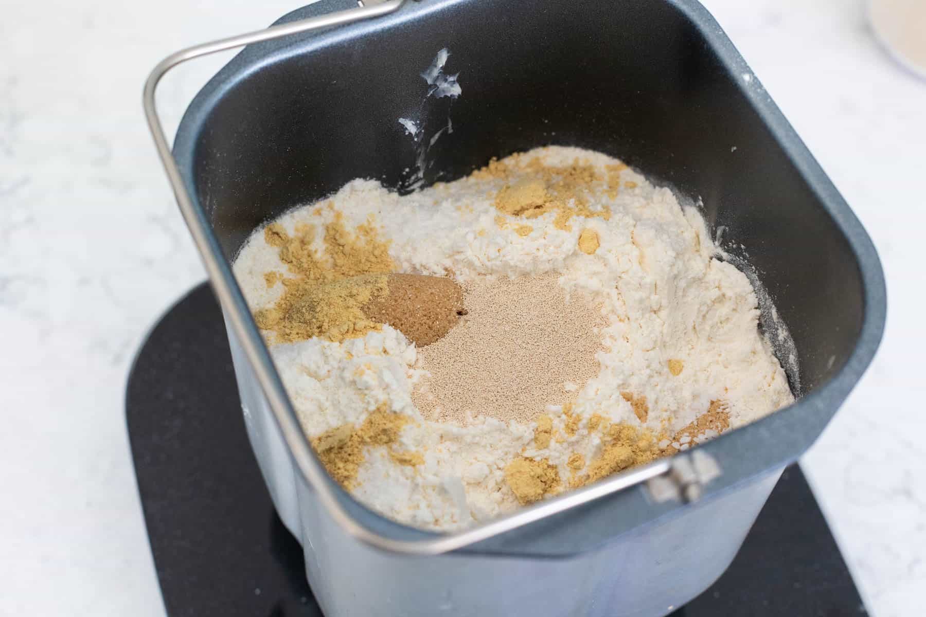 A bread maker pan filled with flour, ground ginger, brown sugar, and dry yeast piled on top, ready to mix for gingerbread bread.
