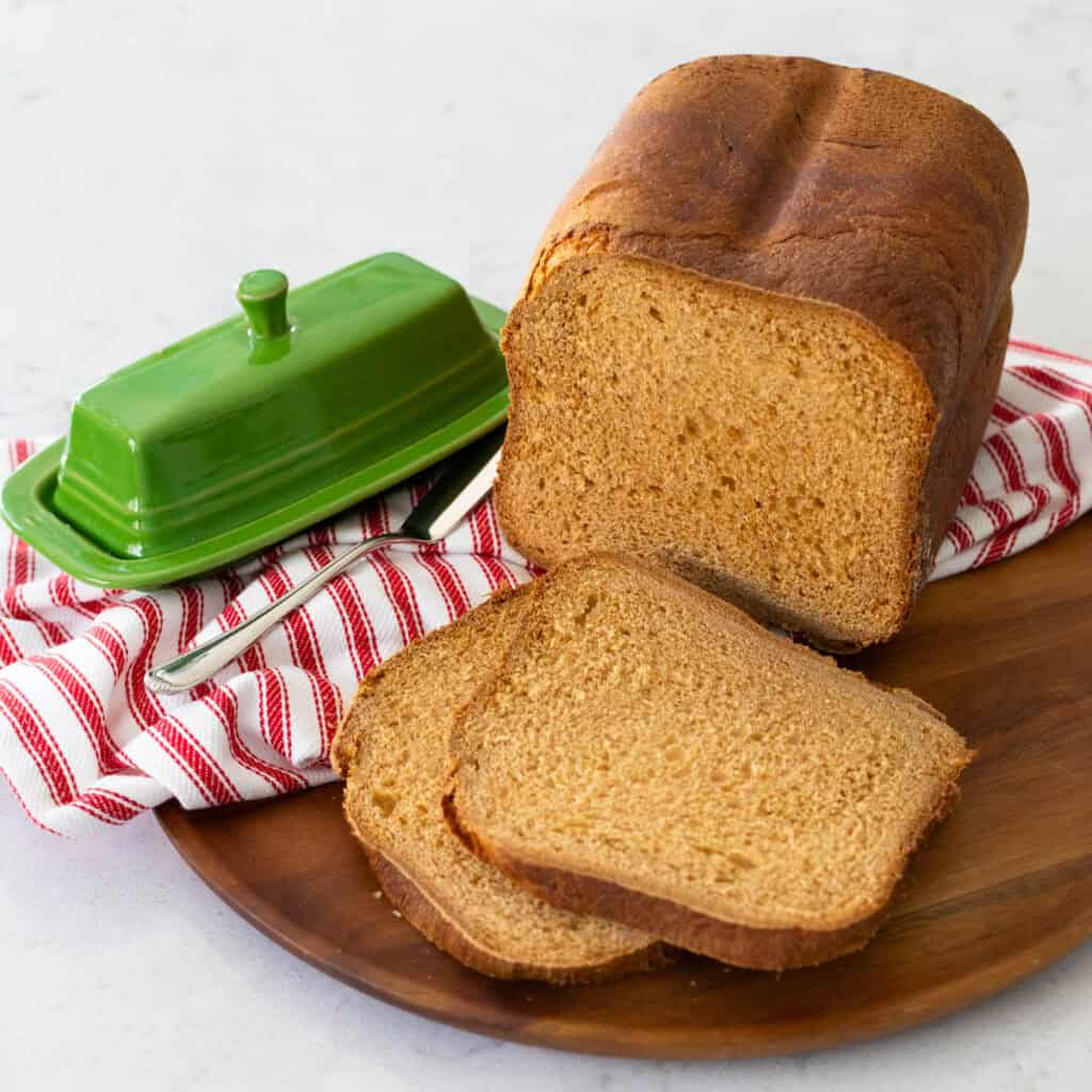 A loaf of bread machine gingerbread loaf has been sliced and served on a wooden platter with a green butter dish and a red and white striped napkin.