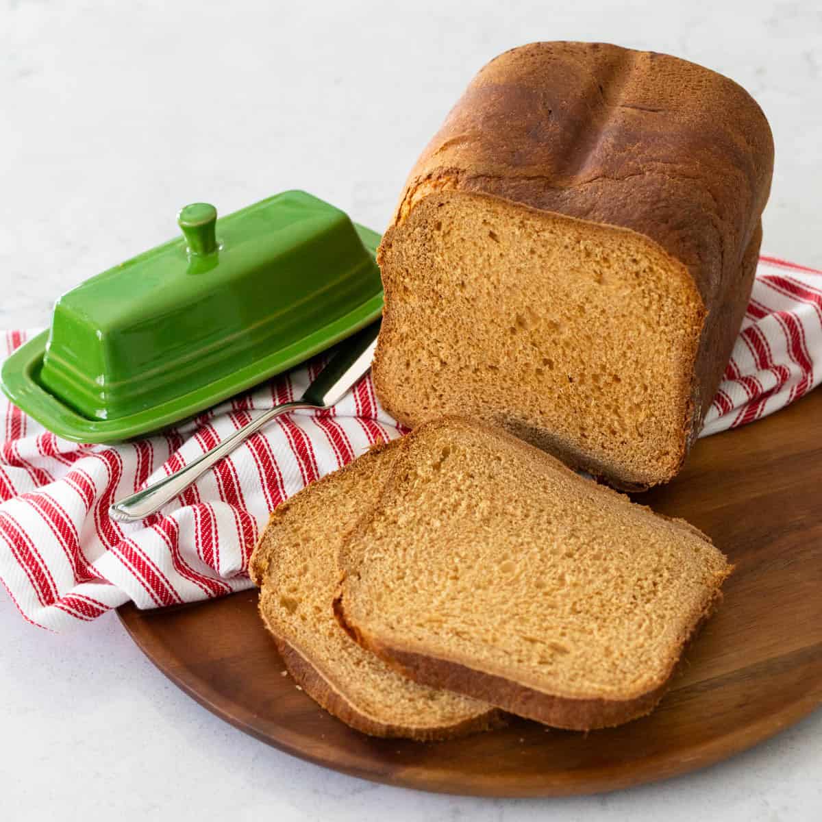 A loaf of bread machine gingerbread loaf has been sliced and served on a wooden platter with a green butter dish and a red and white striped napkin.