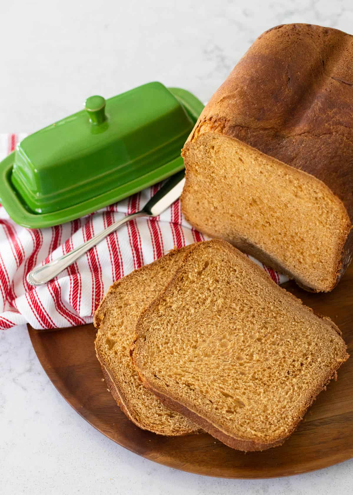A loaf of bread machine gingerbread loaf has been sliced and served on a wooden platter with a green butter dish and a red and white striped napkin.