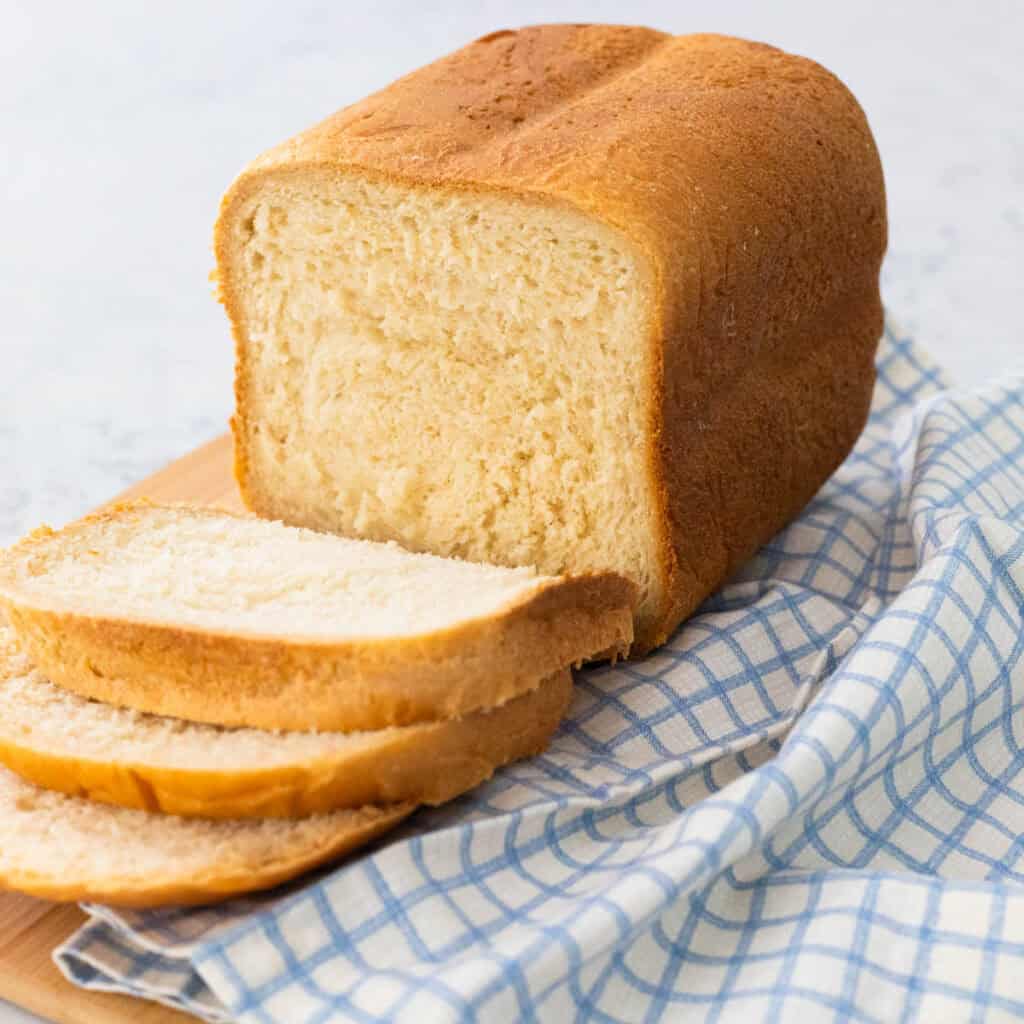 A loaf of potato bread sits on a cutting board, several slices have been laid out in front to show the texture. a blue checked napkin is draped on the side.
