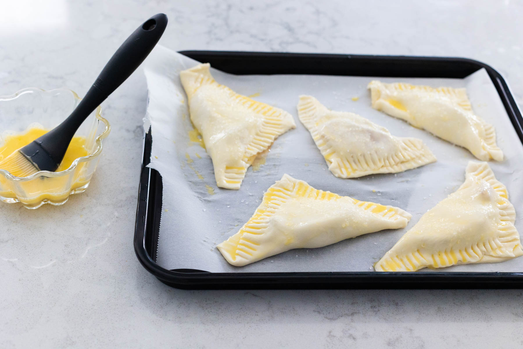 The turnovers on an air fryer pan with a cup of egg wash and a pastry brush on the side.