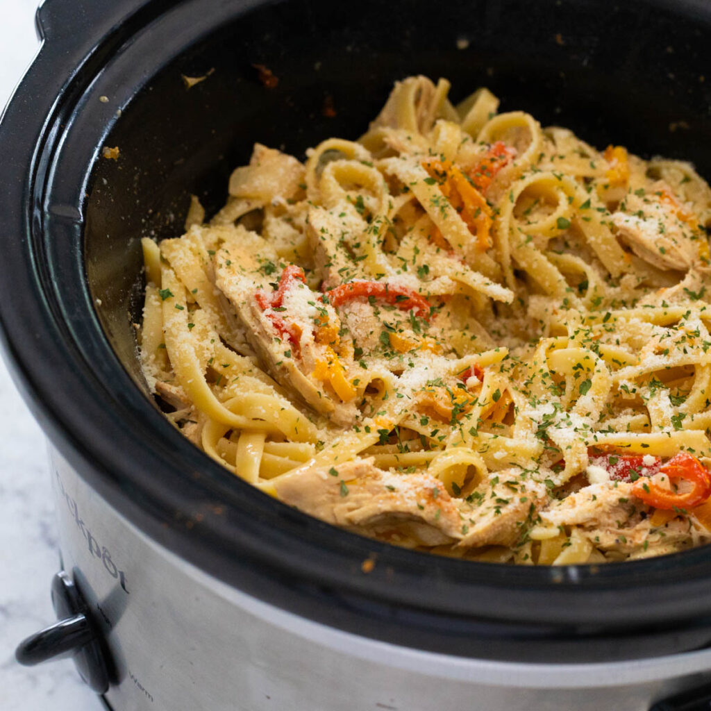 The slowcooker is filled with long noodles, chunks of chicken, bell peppers, and is sprinkled with grated parmesan cheese on top.