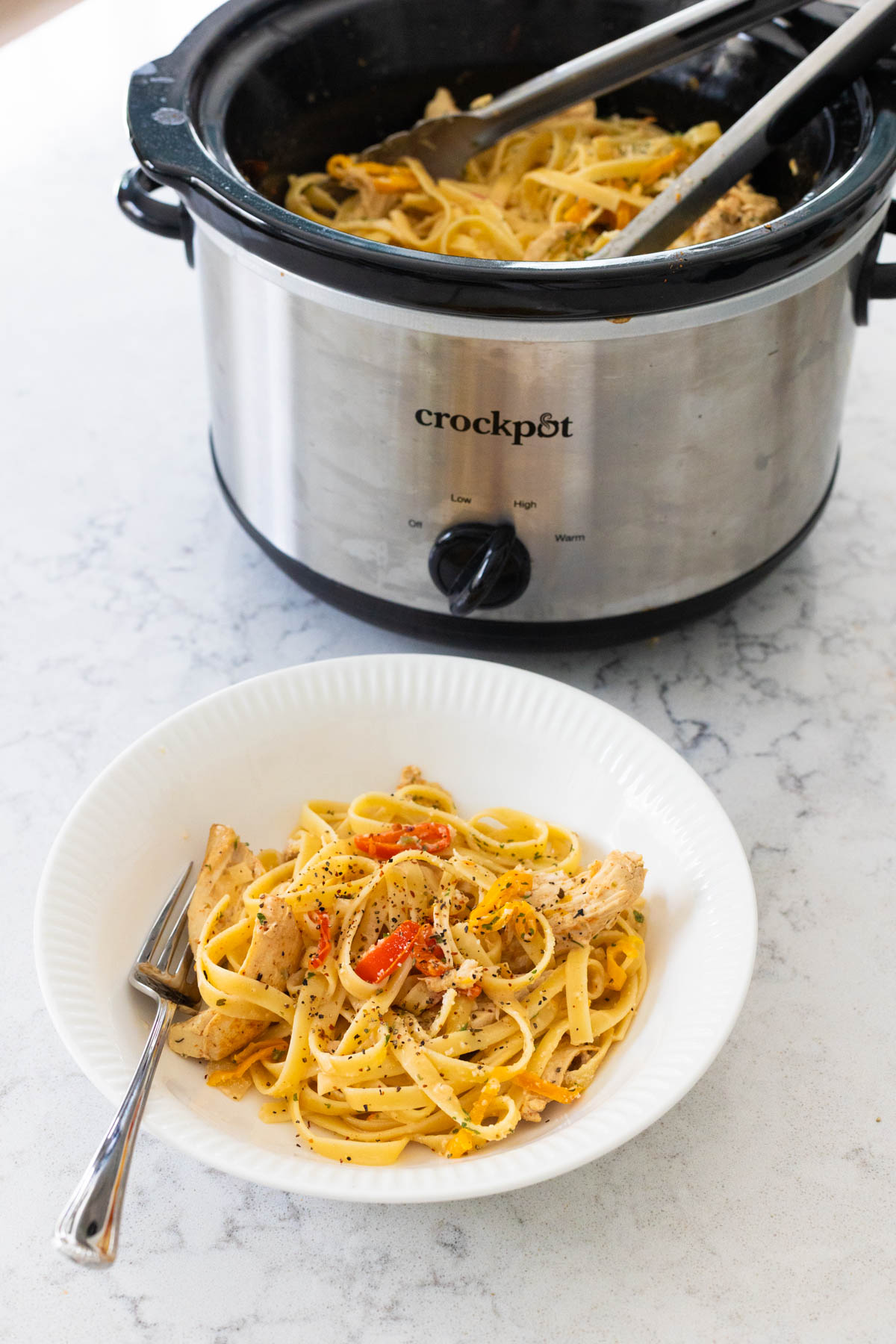 A bowl of creamy cajun chicken pasta with a fork sits in front of the crockpot.