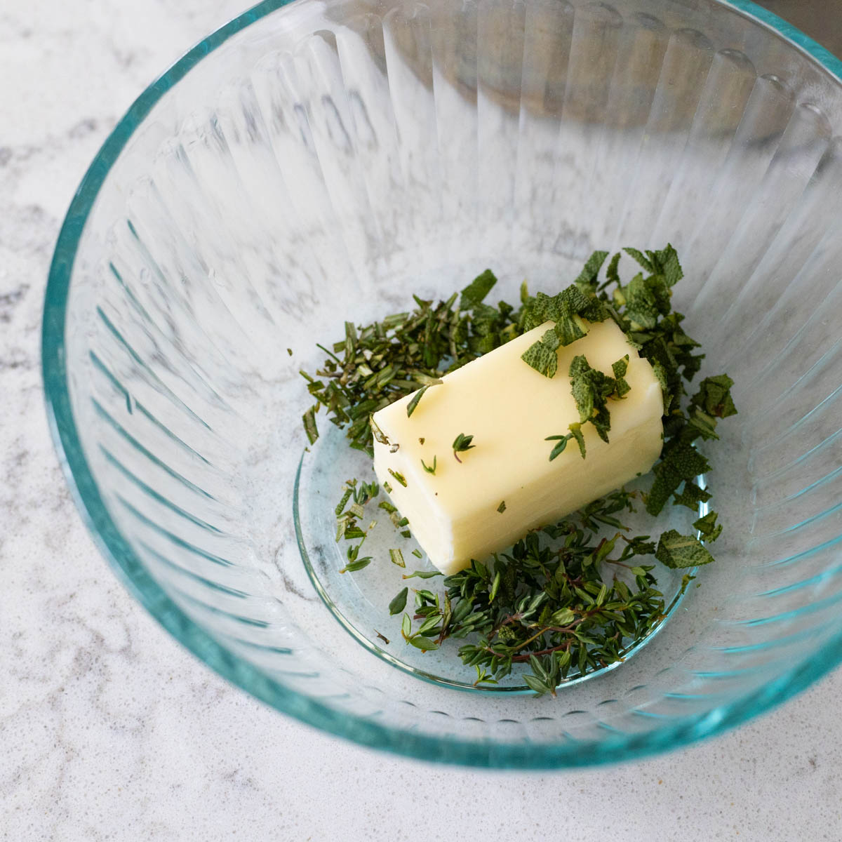 A section of butter has fresh chopped herbs sprinkled over it in a mixing bowl.