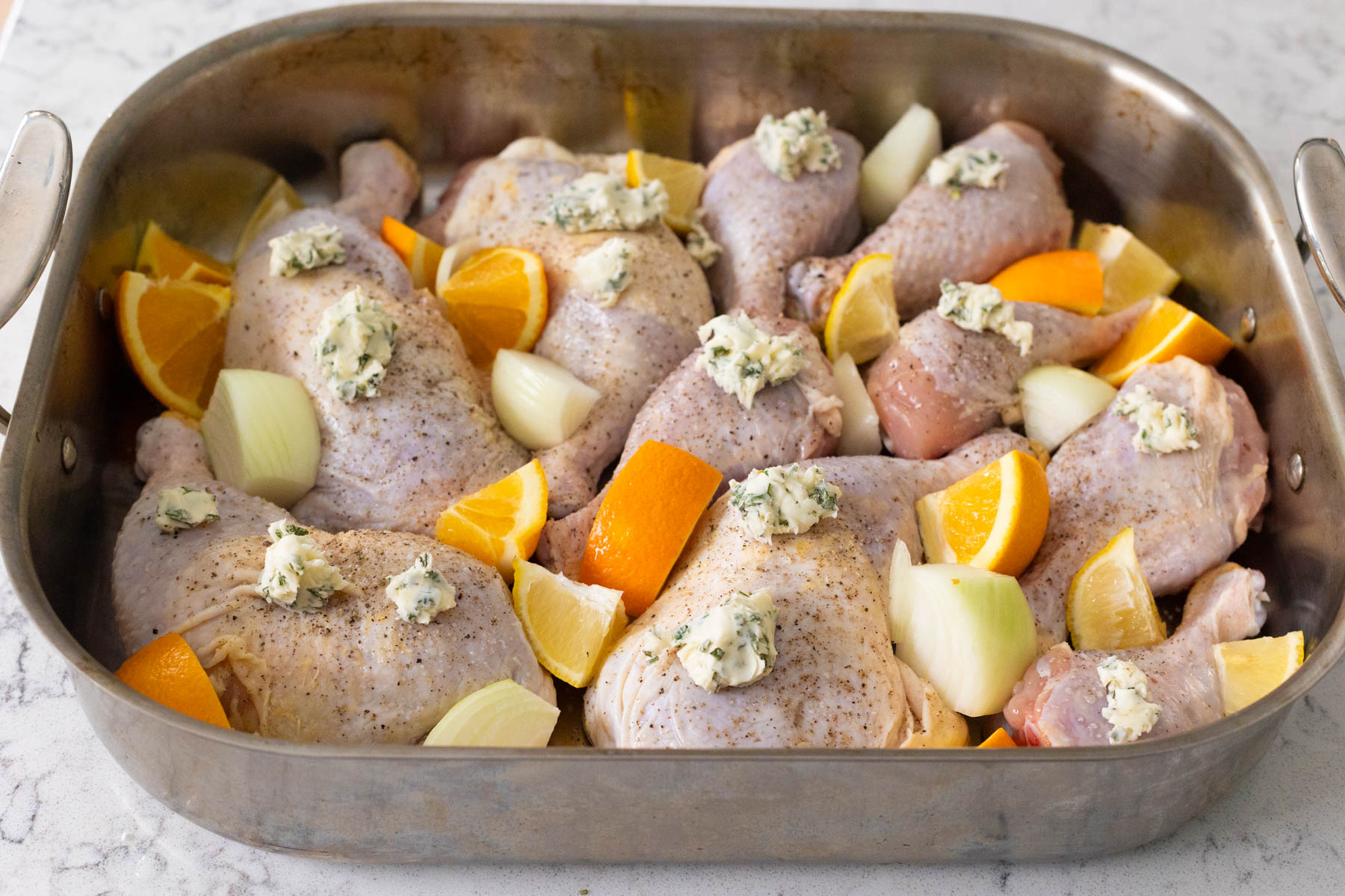 The chicken leg quarters are in a roasting pan with orange, onion, and lemon segments and herb butter dotting the tops.
