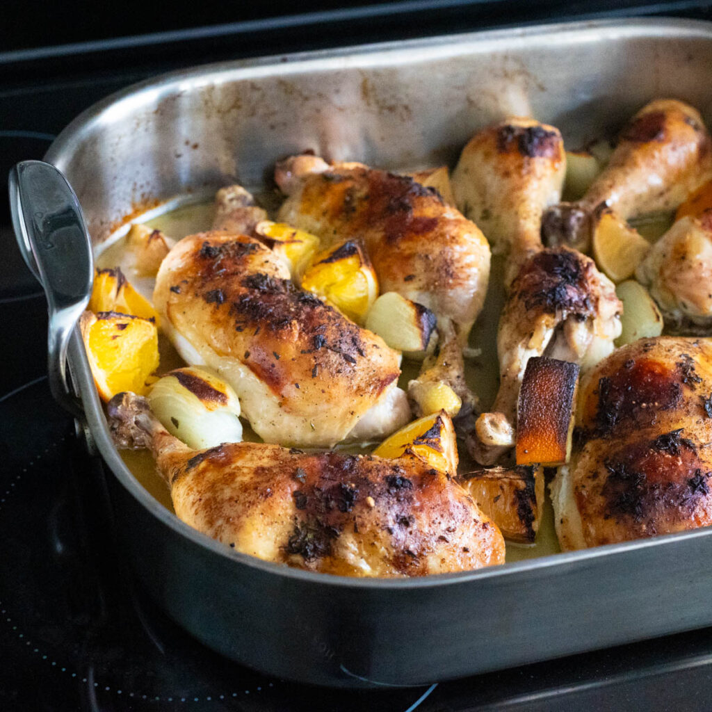 A roasting pan filled with chicken leg quarters with onion, lemon, and orange pieces.