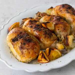 A white platter has 4 chicken leg quarters that have been roasted in the oven to a golden brown. There are roasted orange and lemon segments on the side.