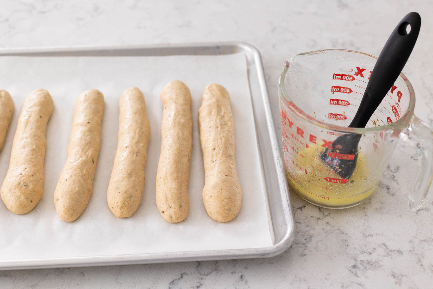The rolled breadsticks are on a baking pan alonside a cup of melted butter that is ready to brush them with a pastry brush.