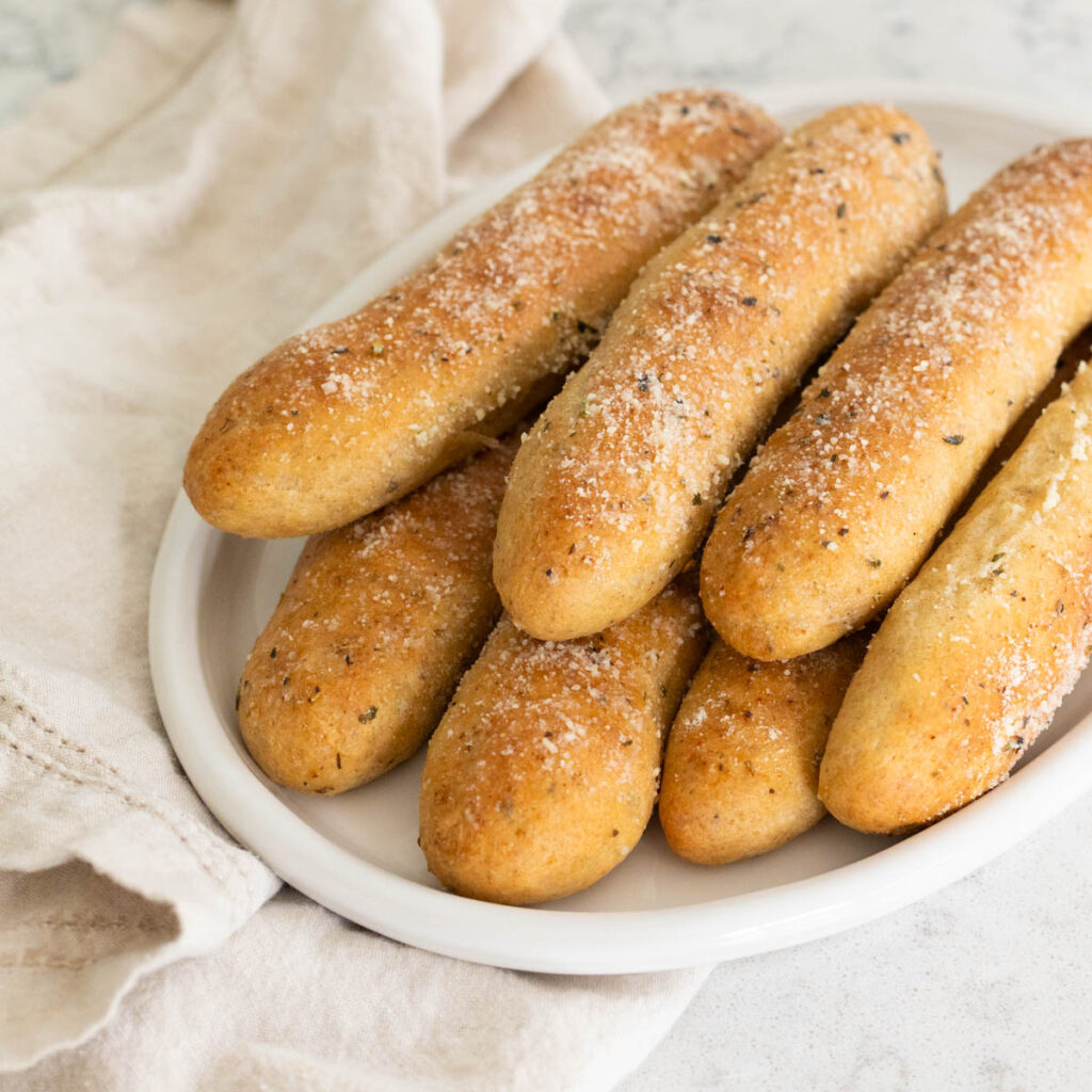 A white platter has a pile of fluffy whole wheat breadsticks dusted with grated parmesan cheese over the top.