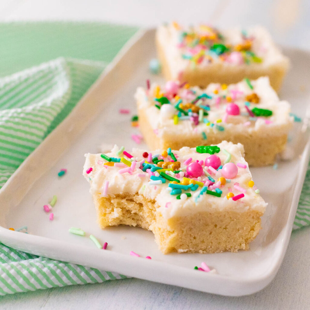 A white platter has 3 sugar cookie bars with white frosting and colorful sprinkles on top.