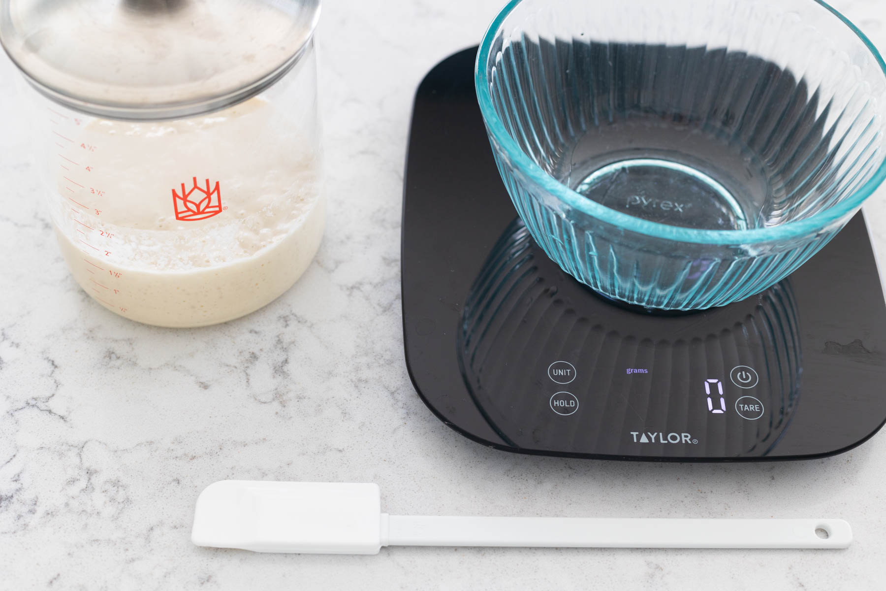 An empty mixing bowl sits on a food scale. A jar of sourdough starter sits next to a thin spatula.