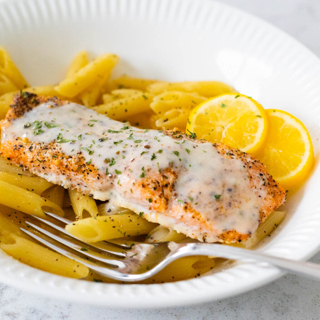 A salmon filet rests on a bowl of pasta. It has been coated in lemon cream sauce with fresh lemon slices on the side.