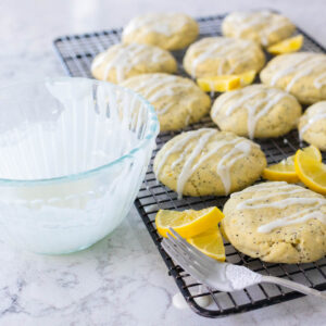 A mixing bowl filled with lemon glaze sits next to a rack of cookies. There's a fork on the side used to drizzle the glaze over the top.