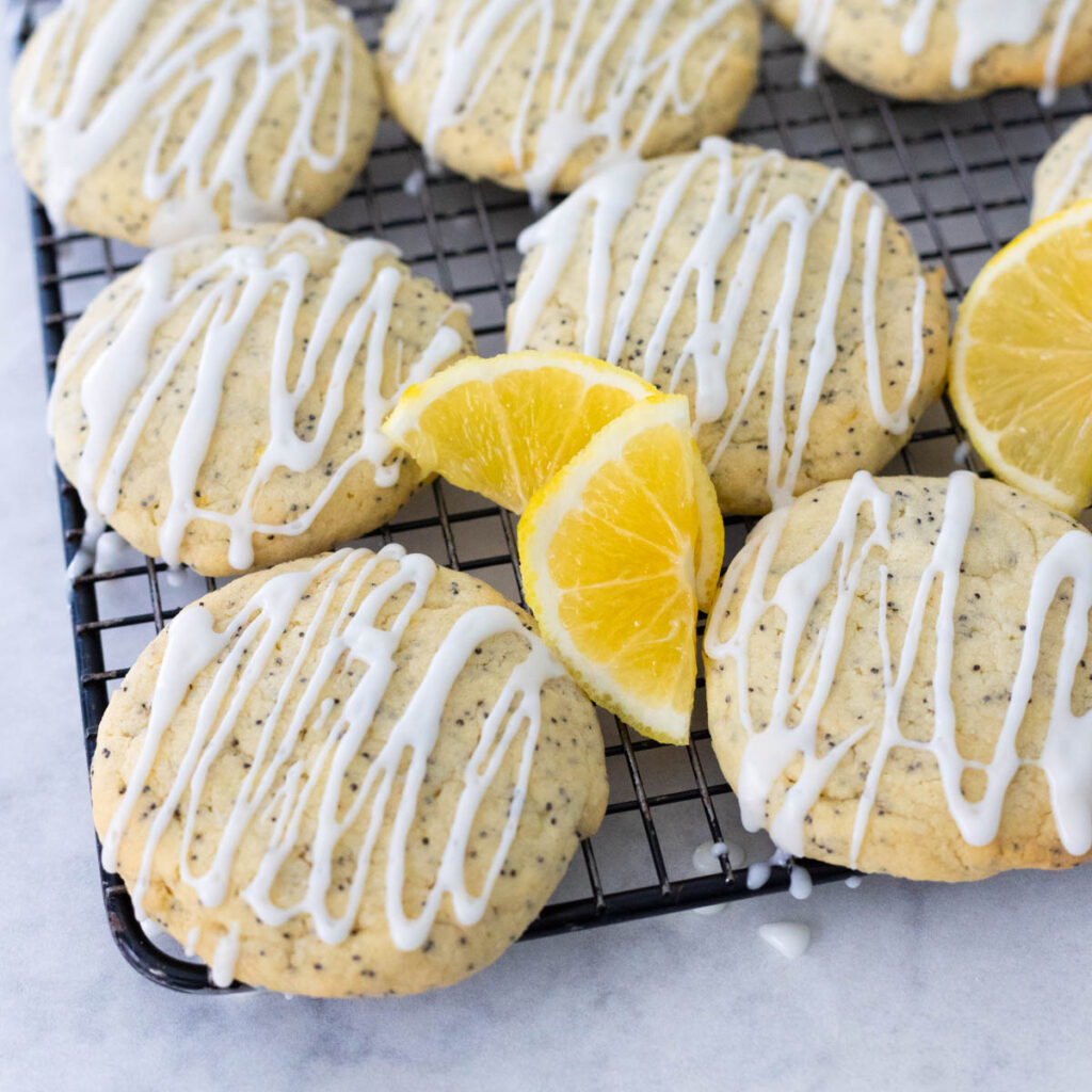 The lemon cookies are cooling on a baker's rack, they have a lemon icing drizzle over the top and fresh lemon slices on the side.