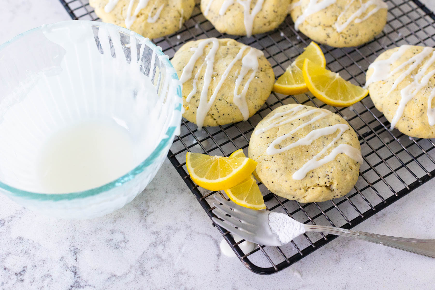 The cookies are on a wire rack. A bowl of lemon glaze is off to the side. A fork used for drizzling the icing rests on the side.