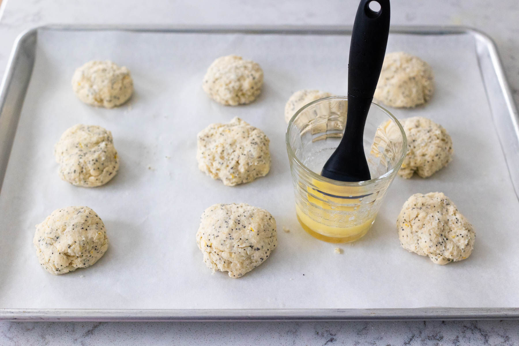 The scones are on a baking pan. A cup of melted butter has a pastry brush dipped in it to top the scones.