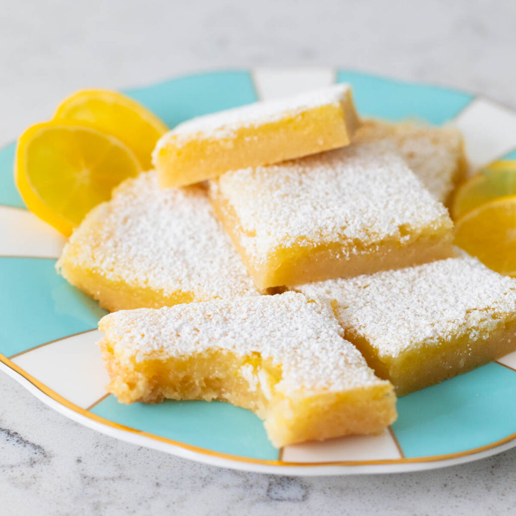 A plate of lemon bars has one square in front that has a bite taken out of it.
