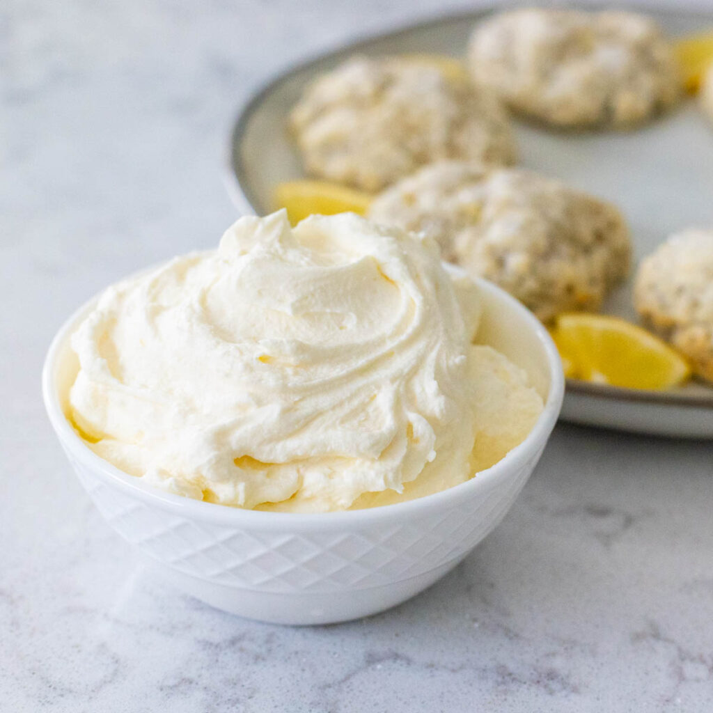 A white bowl is filled with whipped clotted cream and sits in front of a platter of scones.