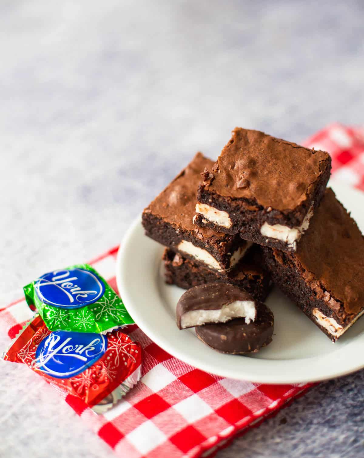 A white plate is stacked with peppermint brownies. Peppermint patty candies are off to the side.