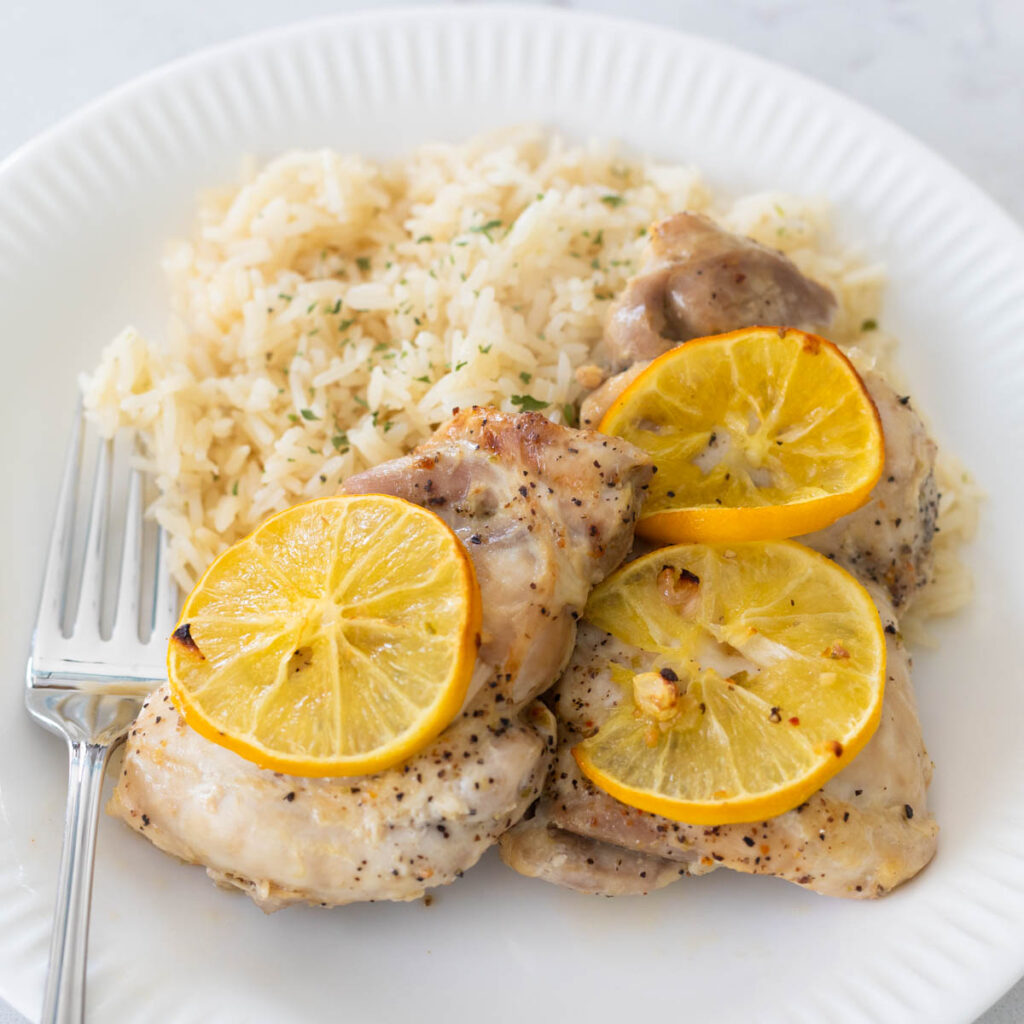 A plate is filled with rice pilaf and Meyer lemon roasted chicken thighs that have sliced lemons on top.