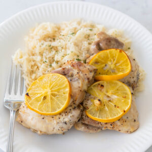 A plate is filled with rice pilaf and Meyer lemon roasted chicken thighs that have sliced lemons on top.