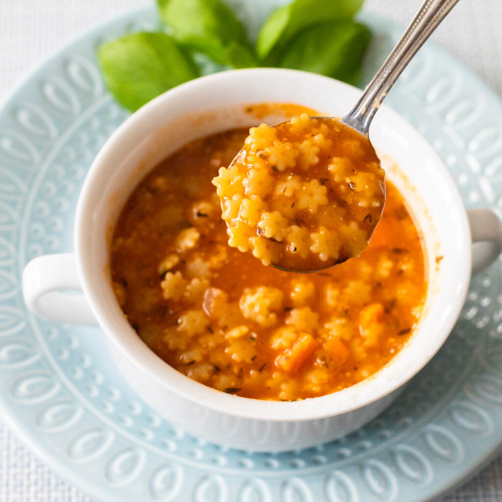 A bowl of tomato-based pastina soup has a spoon lifting out a bite so you can see the star-shaped pasta.