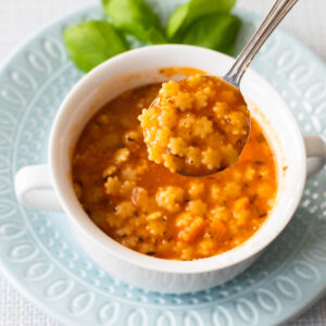 A bowl of tomato-based pastina soup has a spoon lifting out a bite so you can see the star-shaped pasta.
