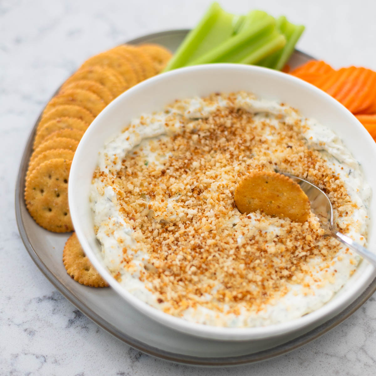 A big bowl of creamy pickle dip has toasted bread crumbs and chopped fresh chives sprinkled over the top. There's a cracker being dunked in the dip.