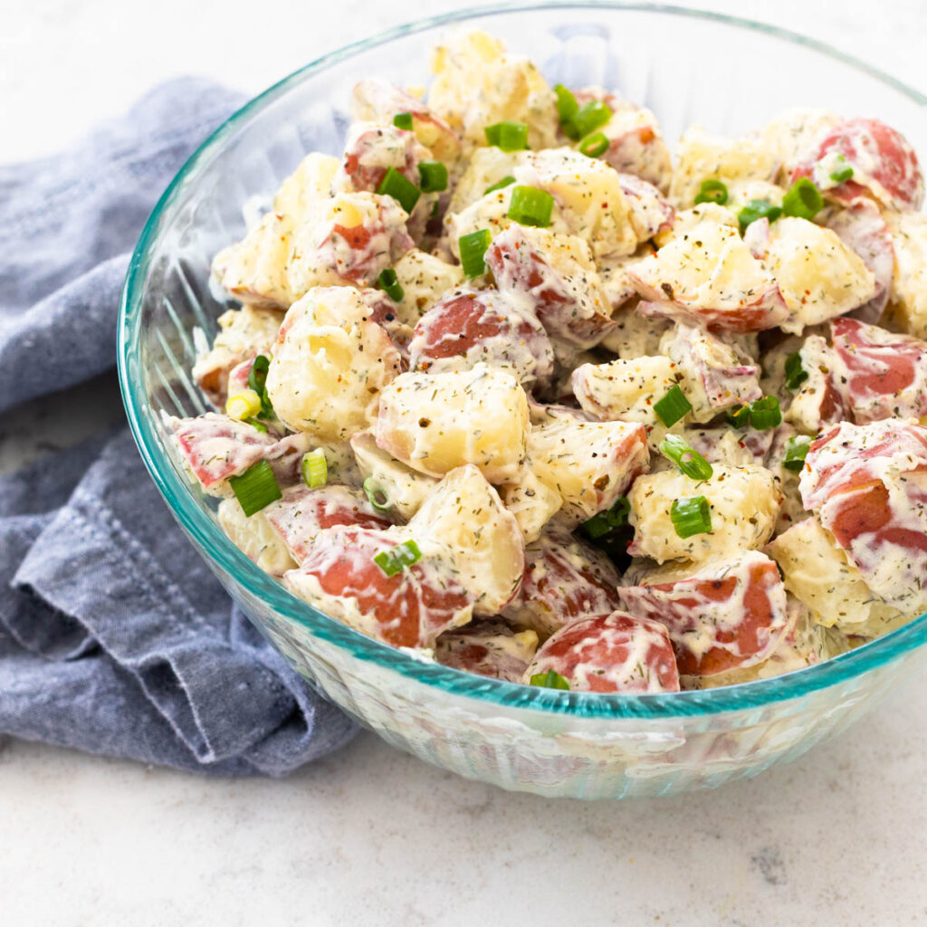 The serving bowl is filled with a creamy dill potato salad made from red potatoes with green onions sprinkled over the top.
