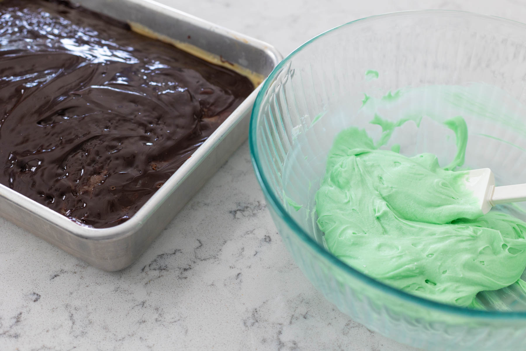 The chilled cake is ready to be frosted with the green frosting in the bowl next to it.