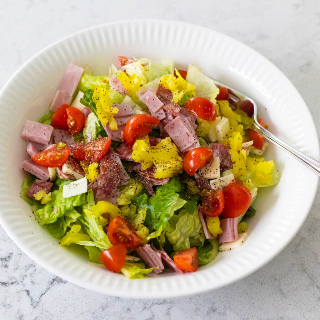 A white bowl is filled with lettuce, tomatoes, and chopped deli meats and cheeses.