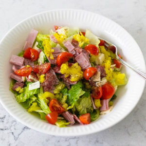 A white bowl is filled with lettuce, tomatoes, and chopped deli meats and cheeses.
