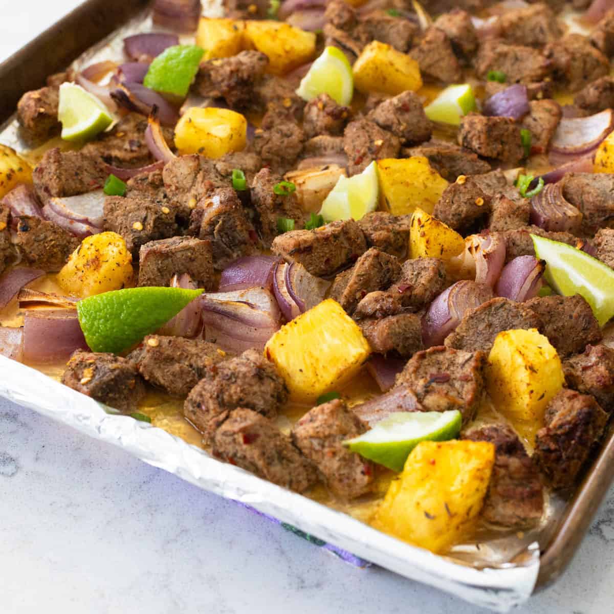 A sheet pan is filled with cooked chunks of jerk pork tenderloin, chunks of fresh pineapple, and lime wedges.