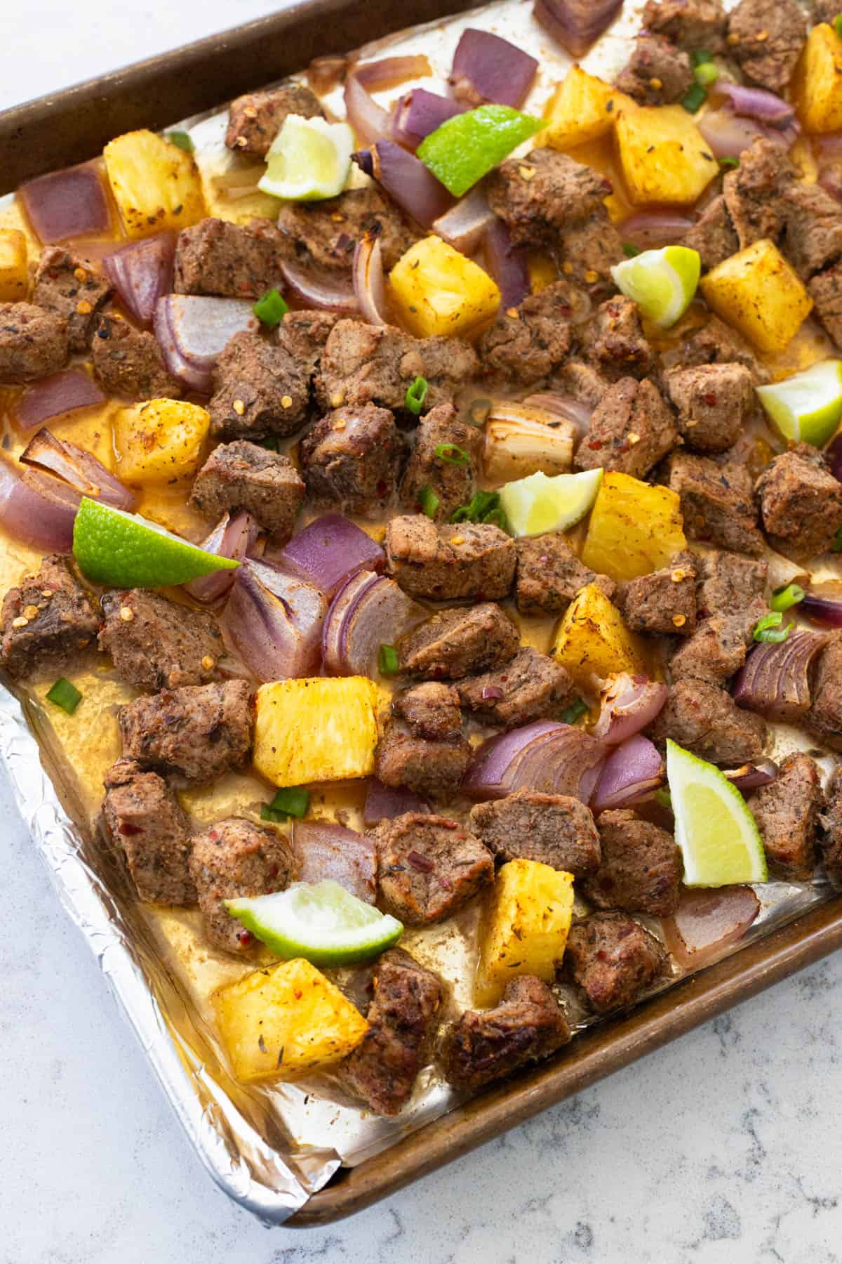 A sheet pan is filled with cooked chunks of jerk pork tenderloin, chunks of fresh pineapple, and lime wedges.