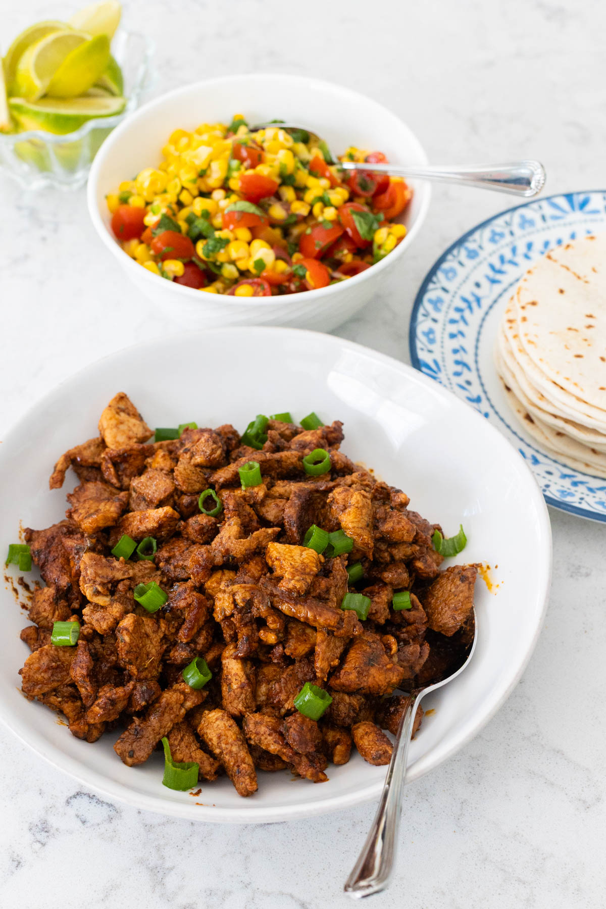 The pork is in a white serving bowl next to a plate of tortillas and a bowl of corn salsa.