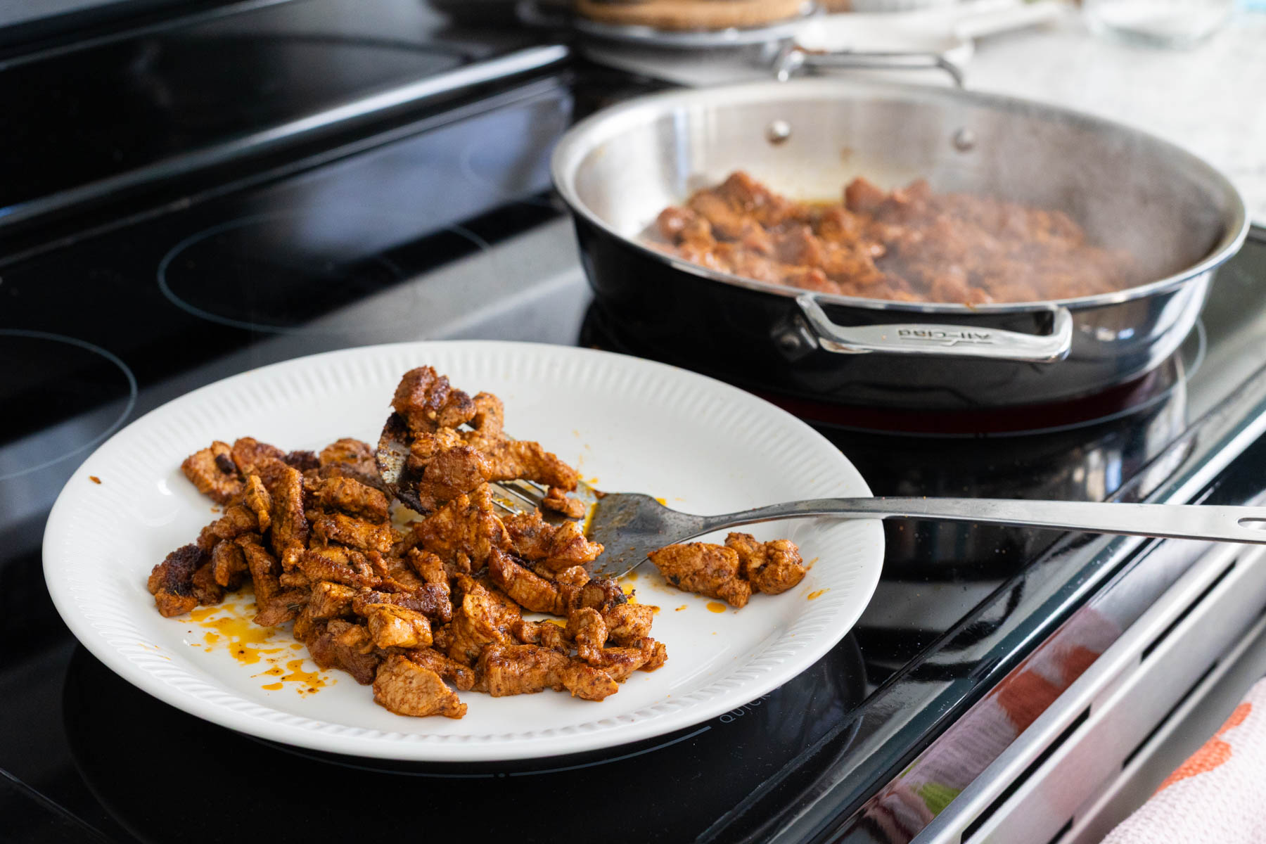 A clean plate has cooked pork on it, the rest of the batch is in the skillet next to it.