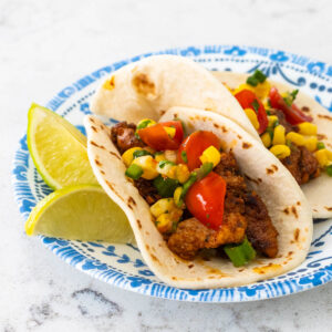 A pork taco with corn salsa on a blue plate, there are lime wedges on the side.